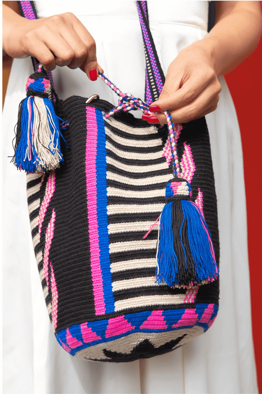 Handwoven Wayuu bag with black, cream, pink, and blue stripes, featuring braided drawstrings and colorful tassels.