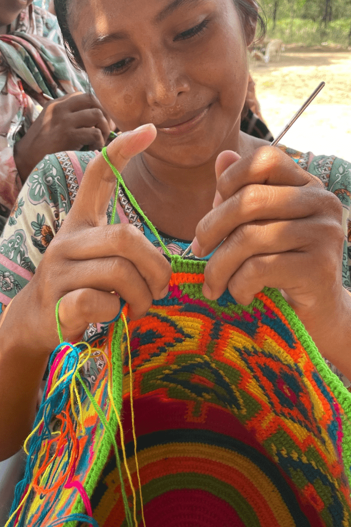 A Wayuu artisan skillfully hand-weaving a traditional mochila bag using vibrant, colorful threads. Her focused expression and delicate craftsmanship highlight the dedication and cultural heritage of the Wayuu people in Colombia.
