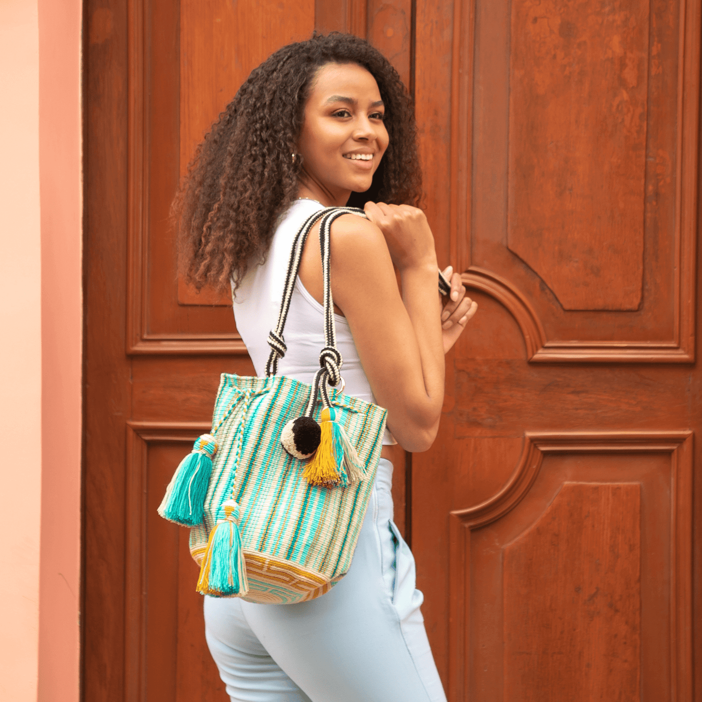A handcrafted Wayuu bag with a vibrant striped pattern in turquoise, white, and yellow. The bag features a single long, braided black-and-white shoulder strap, a drawstring closure, and decorative tassels with a black pompoms accent.