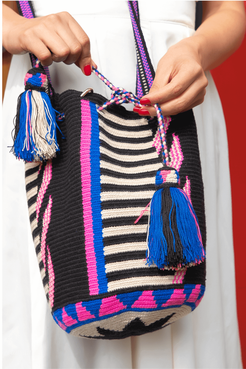 Handwoven Wayuu bag with black, cream, pink, and blue stripes, featuring braided drawstrings and colorful tassels.