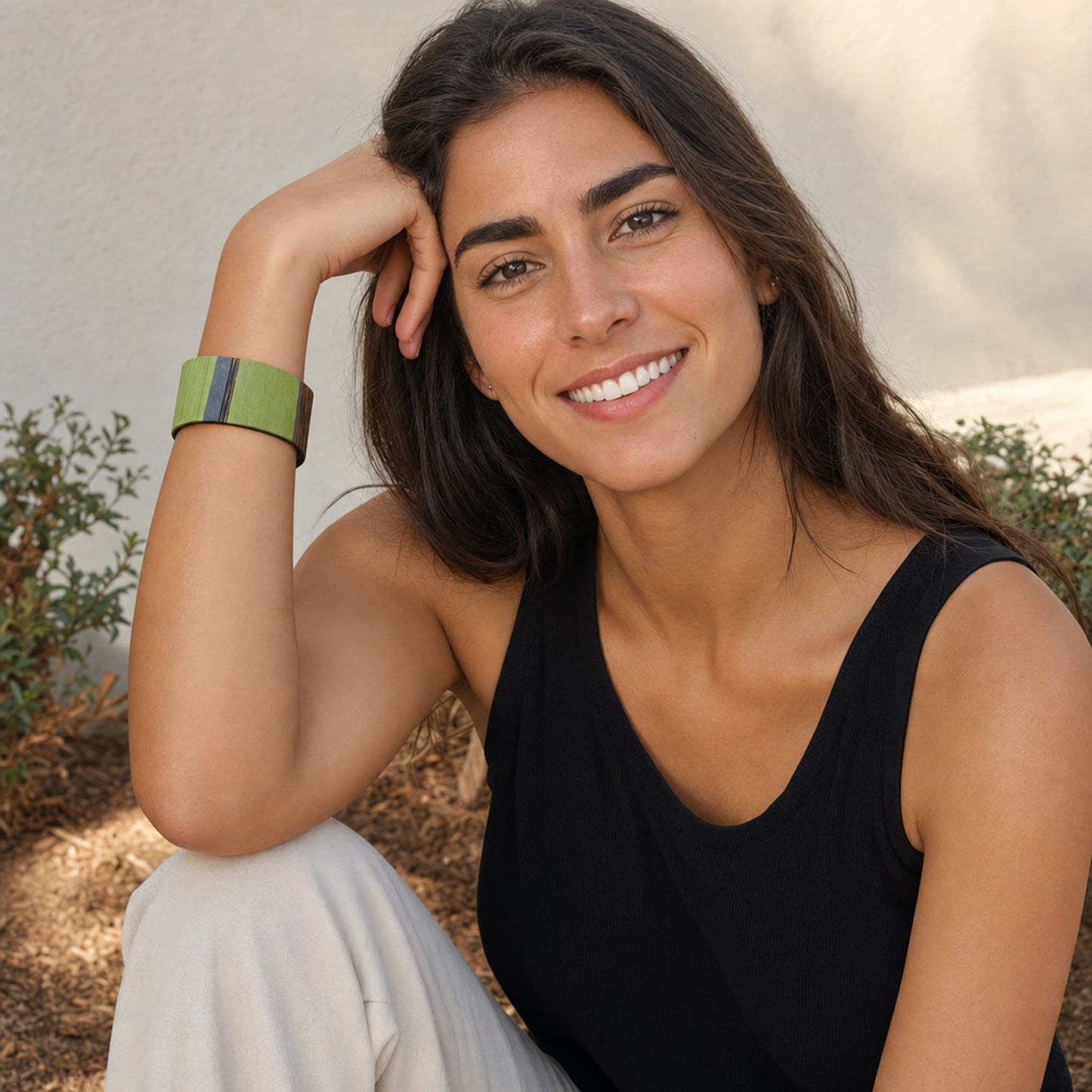 A smiling woman with long dark hair sits outdoors wearing a wide olive green rectangular bracelet with thin blue and purple accent stripes on the outer band. She is wearing a black tank top and light-colored pants, posing against a neutral stucco wall with some plants in the background.