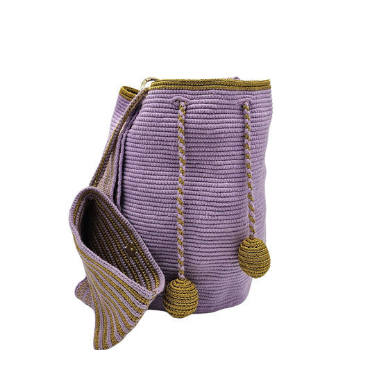 Handcrafted Colombian Wayúu bag in soft lavender with gold braided drawstrings and matching gold pom-poms, featuring a wide shoulder strap with lavender and gold striped detailing.
