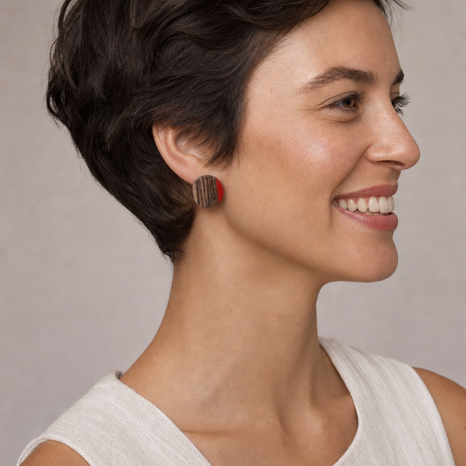 A close-up side profile of a woman with short dark hair wearing a small round wooden earring with a red accent, against a soft neutral background.
