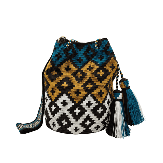 A handmade Wayuu mochila bag with an intricate geometric pattern in black, white, mustard yellow, and teal blue. The cylindrical bag has a woven shoulder strap and a drawstring closure adorned with matching tassels.