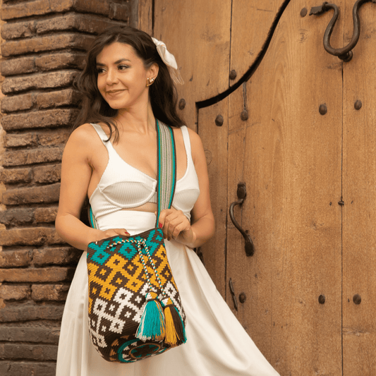 A handmade Wayuu mochila bag with an intricate geometric pattern in black, white, mustard yellow, and teal blue. The cylindrical bag has a woven shoulder strap and a drawstring closure adorned with matching tassels.